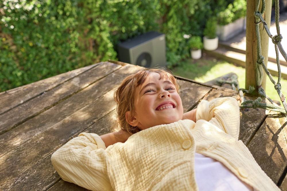 Meisje ligt ontspannen op een houten vlonder; de warmtepomp staat verderop in de tuin