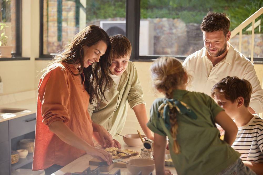 Gezin kookt samen in de keuken; buiten staat de warmtepomp