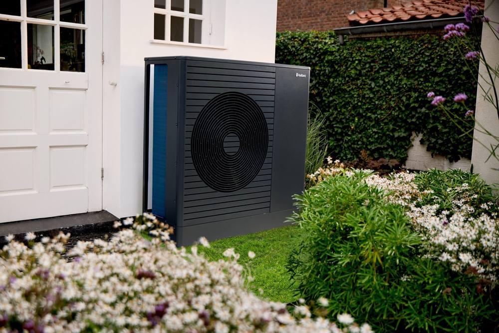 Vaillant aroTHERM pro naast de voordeur van een wit huis, achter een bloeiende border